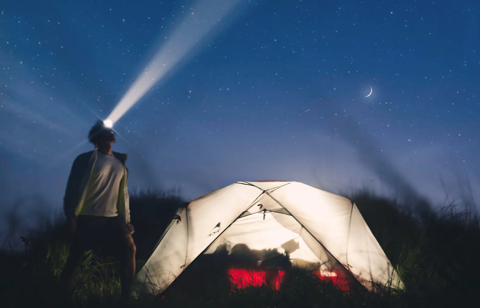 A camper using a headlamp beside a lit tent at night